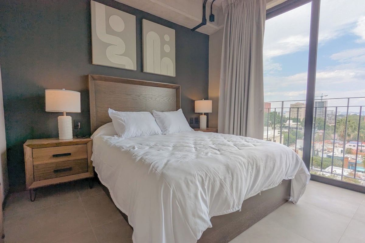 A hotel room with a large bed, two bedside tables with lamps, a wooden headboard, and a view of the city through a balcony.