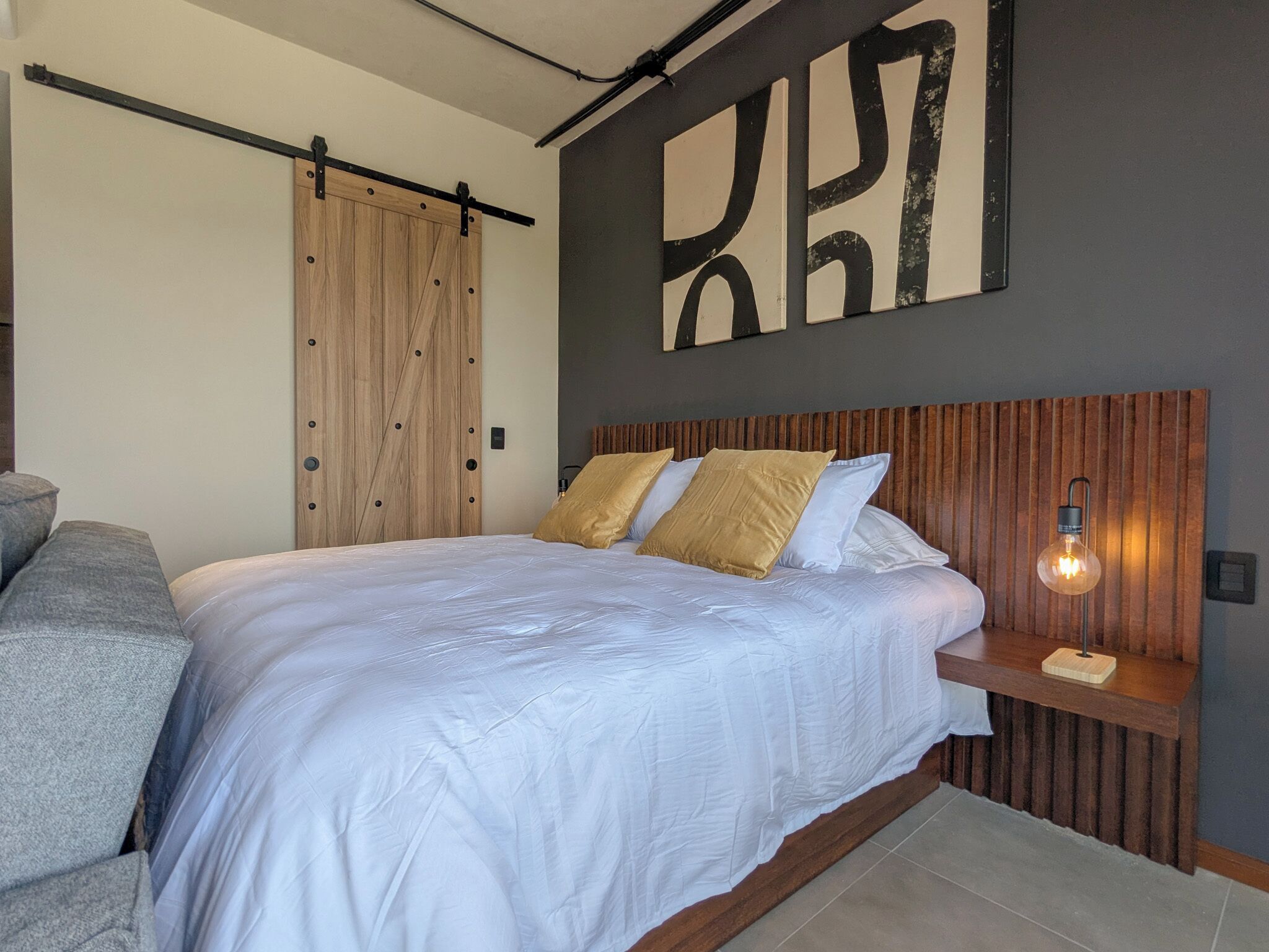 A bedroom with a bed, a wooden wardrobe, a wall-mounted artwork, and a bedside table with a lamp.