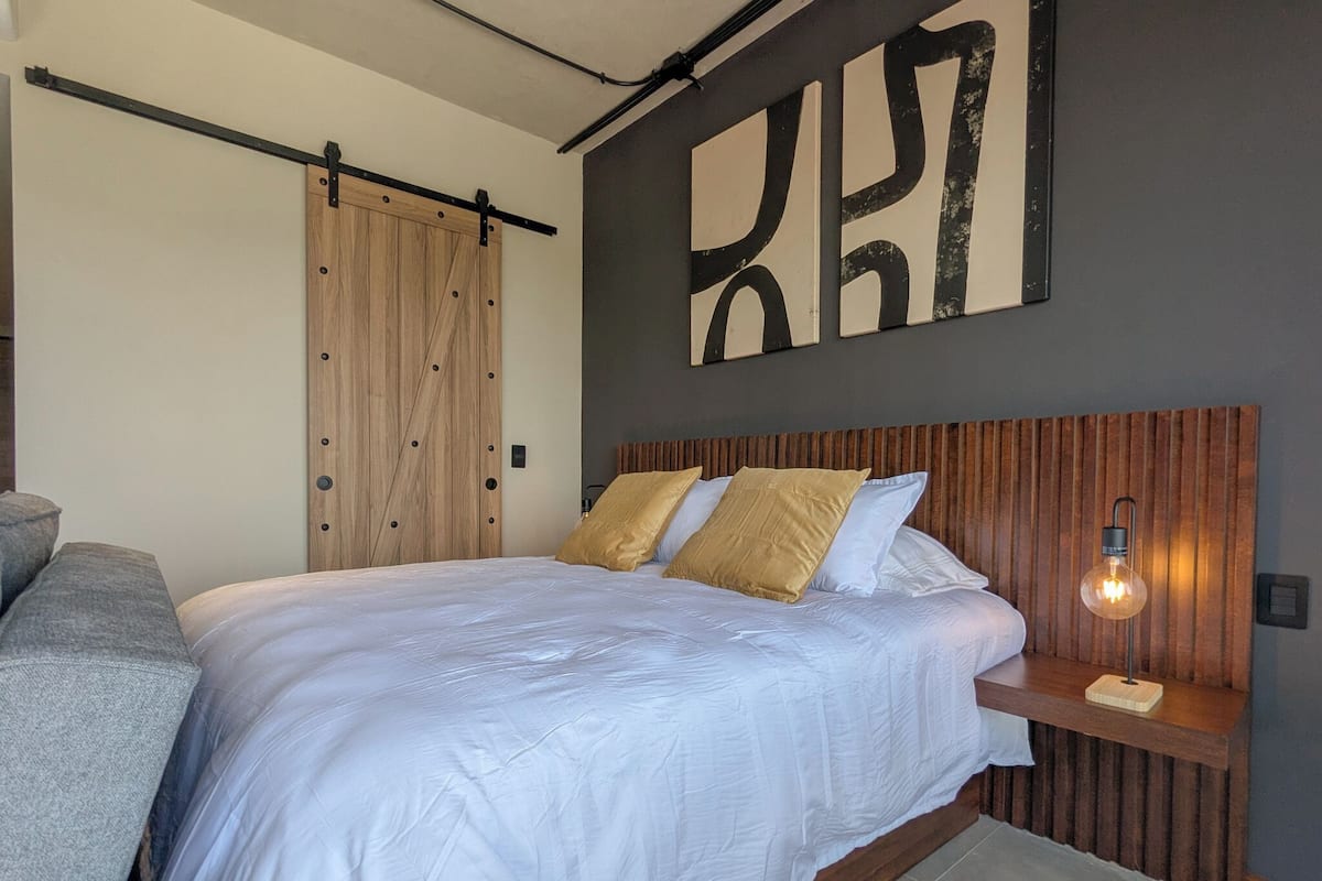 A bedroom with a bed, a wooden wardrobe, a wall-mounted artwork, and a bedside table with a lamp.