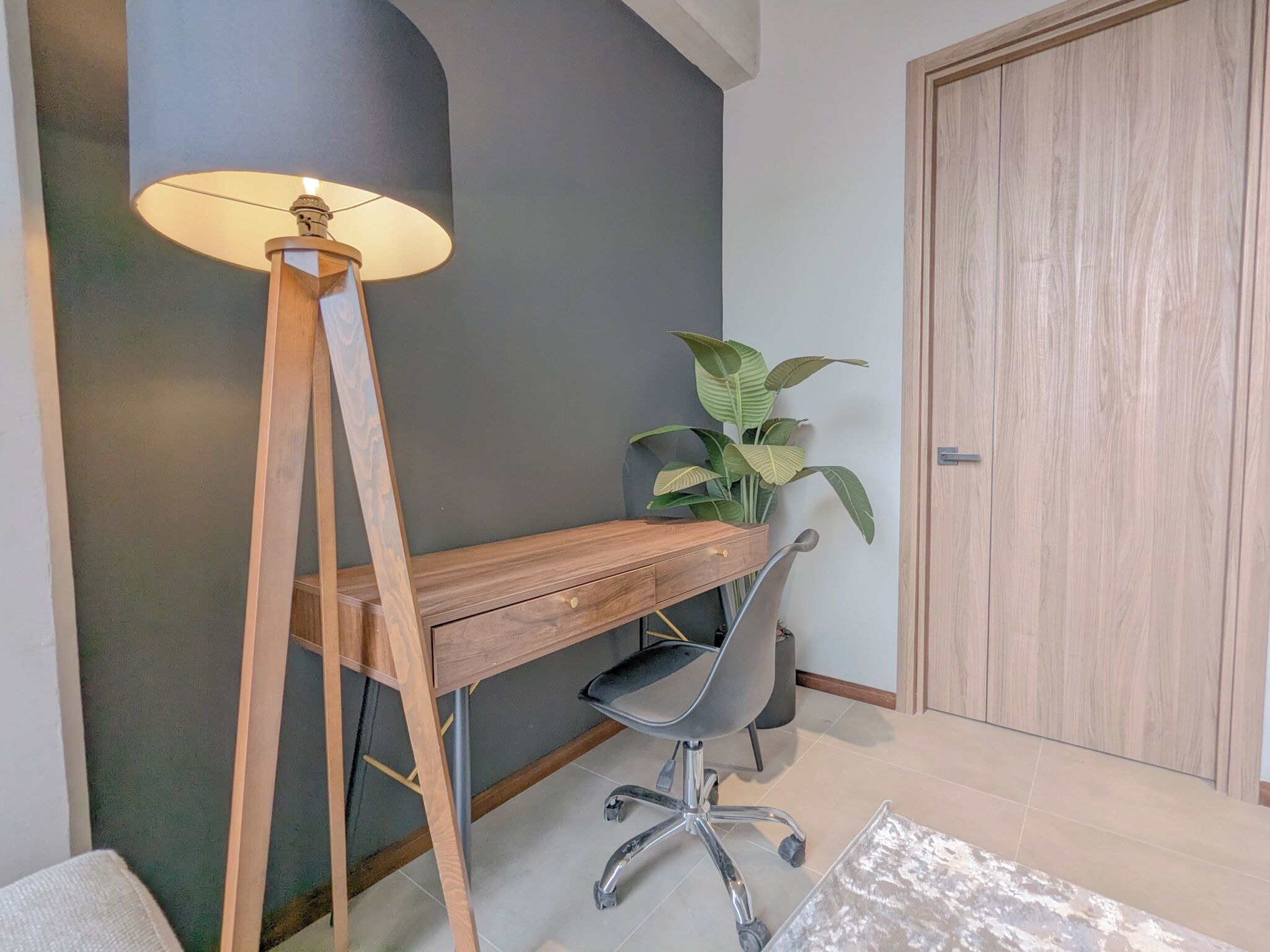 Un escritorio con una lámpara, una silla, una planta en maceta y una puerta en una habitación con paredes grises.