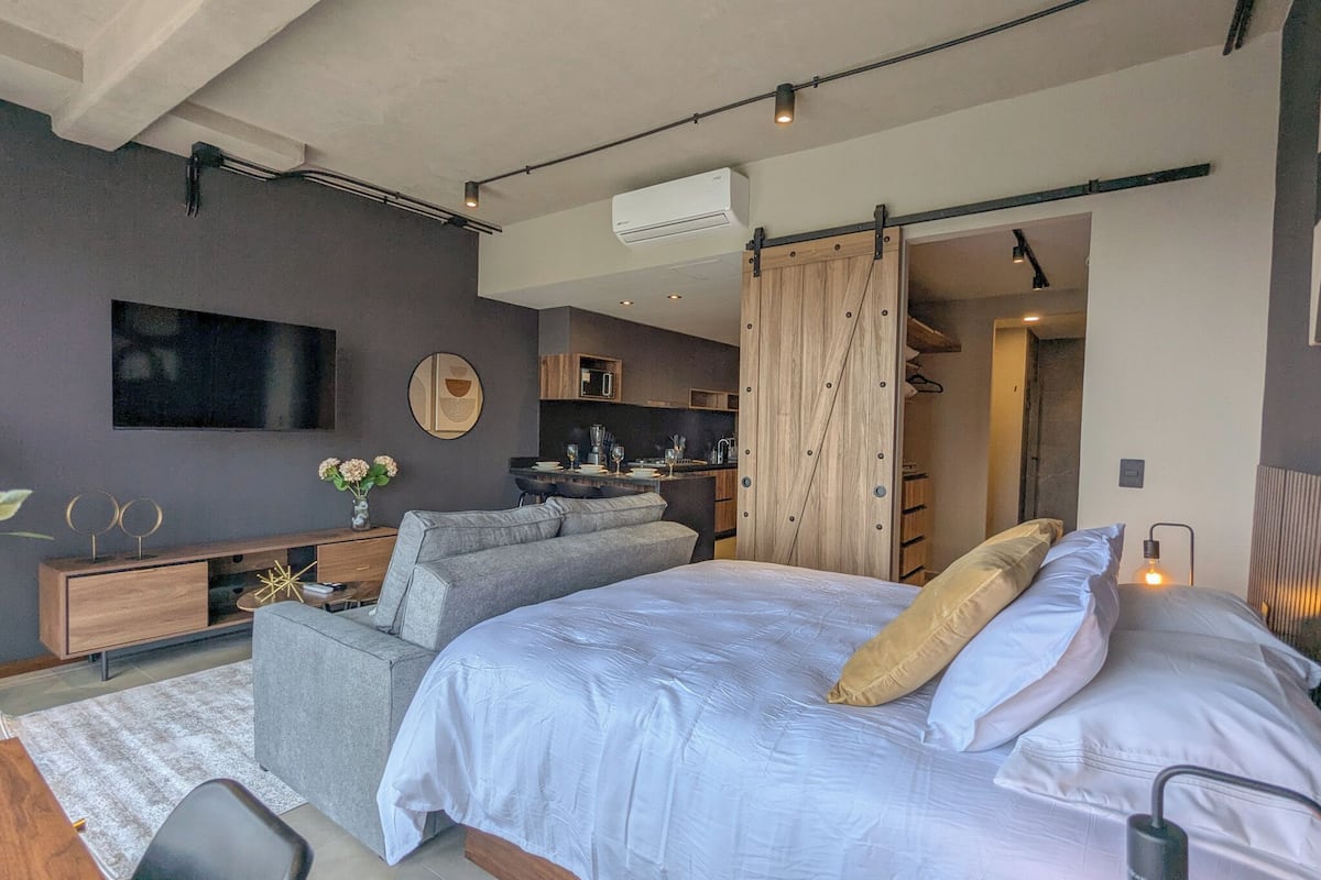 A modern bedroom with a bed, a seating area, a TV, and a kitchen in the background.