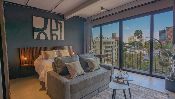 Ein modernes Schlafzimmer mit einem großen Bett, einem grauen Sofa, einem kleinen Tisch und einem großen Fenster mit Blick auf die Stadt.