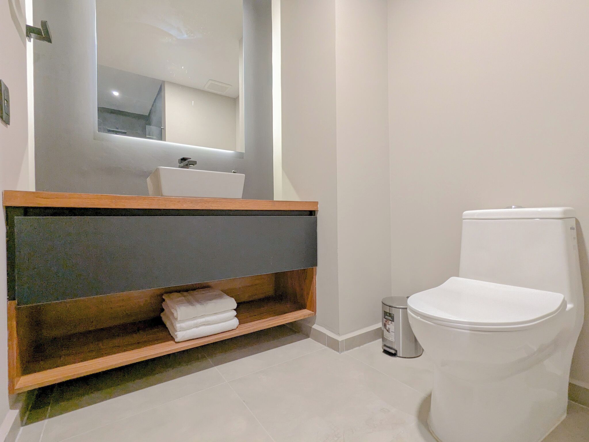 A modern bathroom with a white toilet, a sink, and a mirror above it. There is a shelf with folded towels and a trash bin next to the toilet.