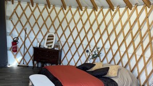 Ein Zimmer mit einem Bett, einem kleinen Hocker, einer Holzkommode und einem Feuerlöscher an der Wand.