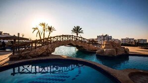 Una piscina amb un pont de pedra, palmeres i edificis moderns al fons.