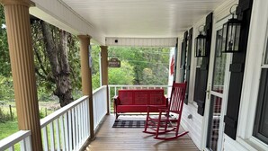 Une véranda couverte avec un banc et des fauteuils à bascule, une balustrade blanche et une vue sur les arbres.