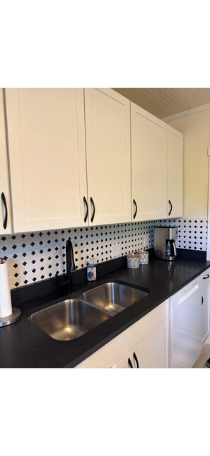 A kitchen with white cabinets, black handles, a black countertop, a stainless steel sink, and a patterned backsplash. - Country Cozy Farmhouse Next to Fairground and Pastures (Sparta)