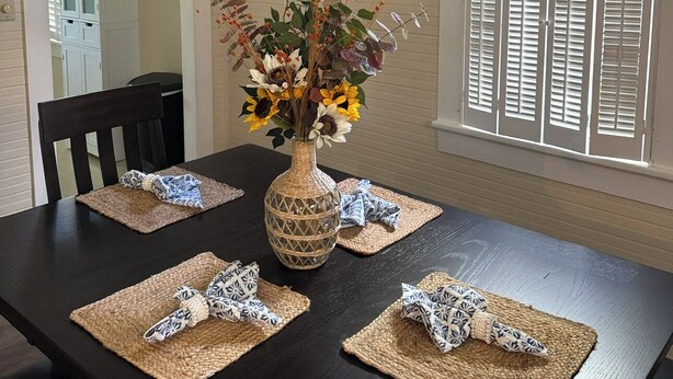 Un coin repas avec une table en bois foncé, des sets de table en fibres tressées, un vase avec des fleurs, un lustre et une fenêtre avec des volets.