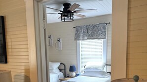 Une chambre à coucher comprenant un lit, un ventilateur de plafond, une fenêtre avec des stores et une table de chevet avec une lampe.