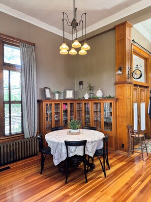 Un comedor con una mesa redonda, sillas, una araña, un aparador de madera con puertas de cristal y una ventana con cortinas.