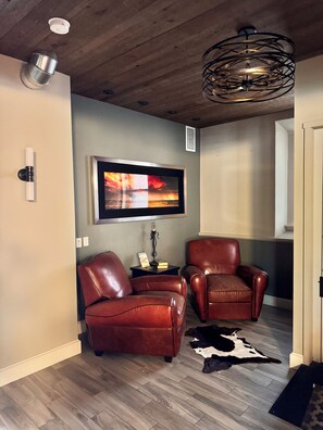 A modern living room with two leather chairs, a mounted TV, a wooden ceiling, and a cowhide rug.