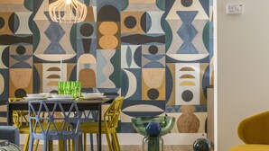 Een eethoek met een wand met een geometrisch patroon, een tafel gedekt met borden en glazen, en stoelen in geel en blauw.