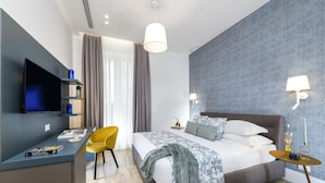 A modern hotel room with a bed, a desk with a flat-screen TV, a yellow chair, and two bedside tables with lamps. - Domus Augustea - Cesare (Roma)