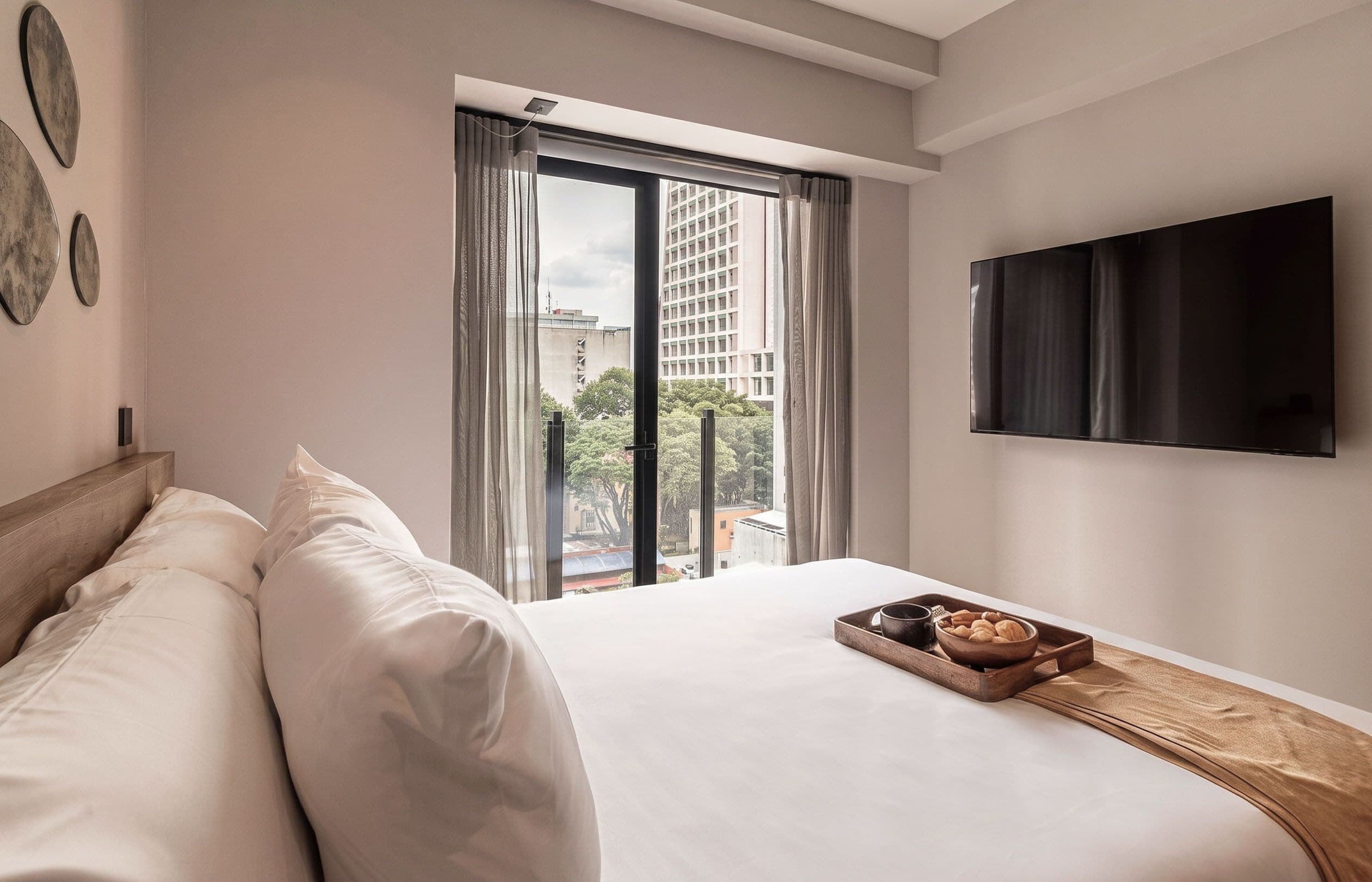 A hotel room with a bed, a TV, a window with curtains, and a tray with bread.