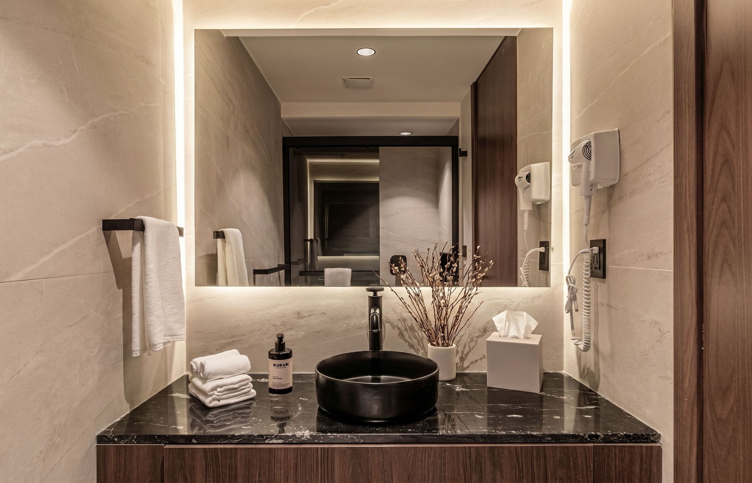 A modern bathroom with a marble countertop, a black vessel sink, a mirror, a towel rack, a plant, and a hand dryer.