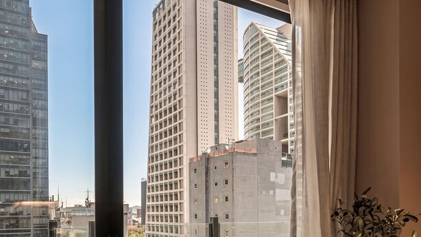 Quarto moderno com uma janela grande, uma mesa redonda com um livro, uma planta em vaso e vista para edifícios da cidade.