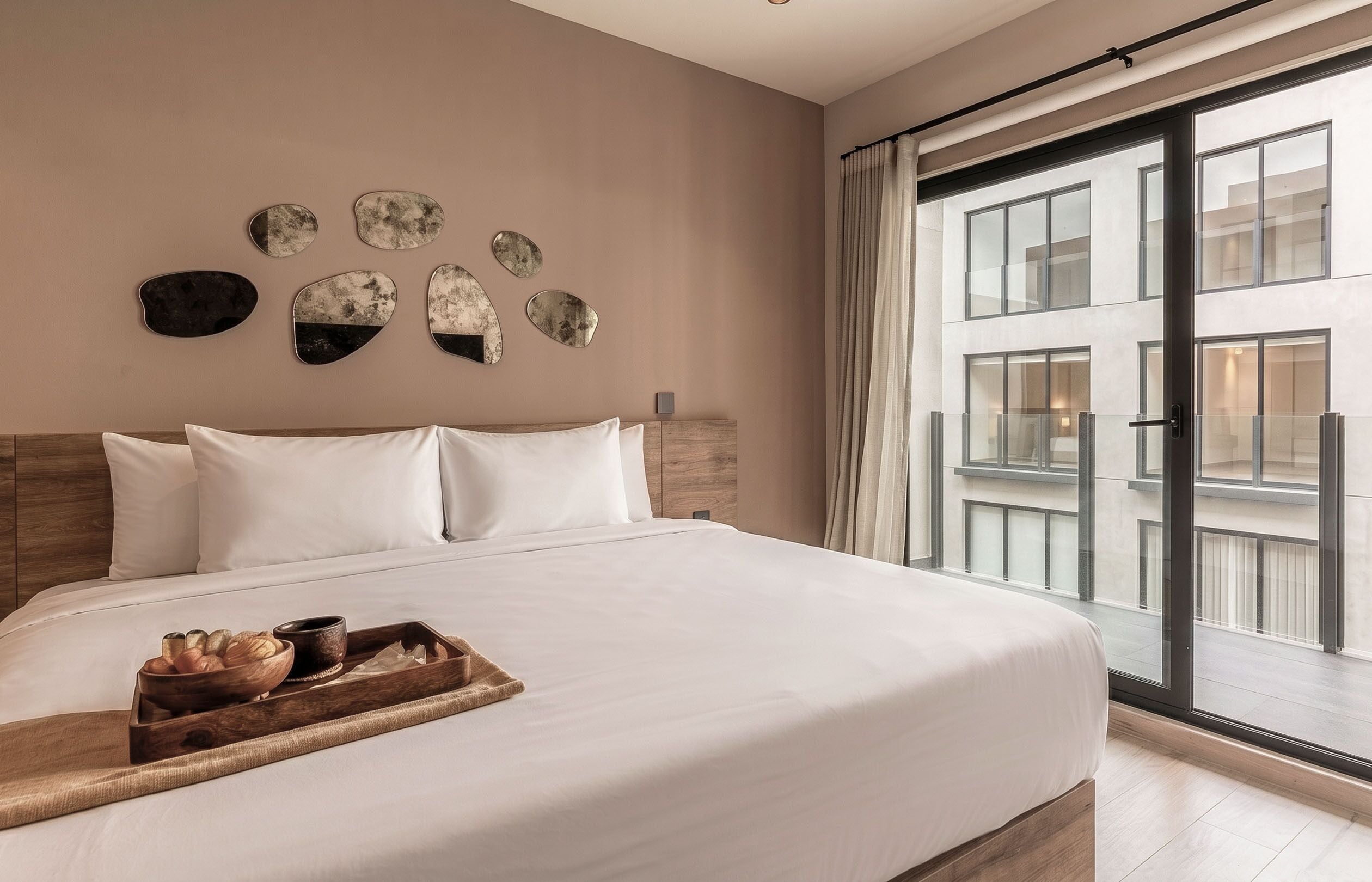 Une chambre d’hôtel avec un grand lit, une tête de lit en bois, un plateau avec du pain et une tasse, et une vue sur un immeuble à travers de grandes fenêtres.