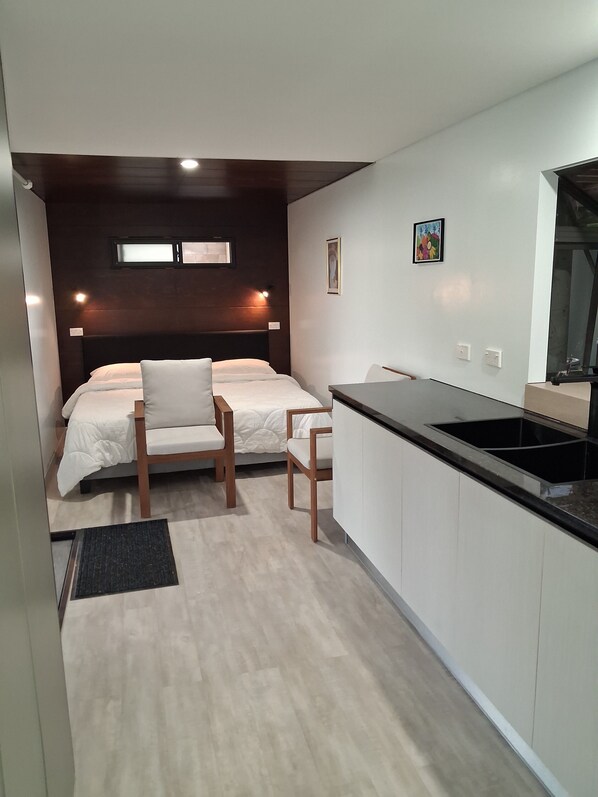 A compact hotel room with a bed, two chairs, a small kitchen area, and a window.