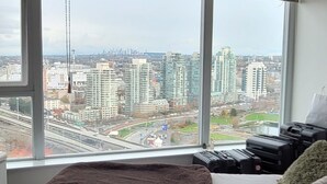 A room with a large window overlooking a cityscape, a bed, and a suitcase.