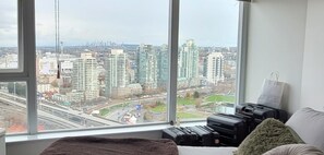 A room with a large window overlooking a cityscape, a bed, and a suitcase. - Pets ok View Suite 3 bed (2 bed + den) 2 bath, 1201 Sqft. Rogers Arena Skytrain (Vancouver)