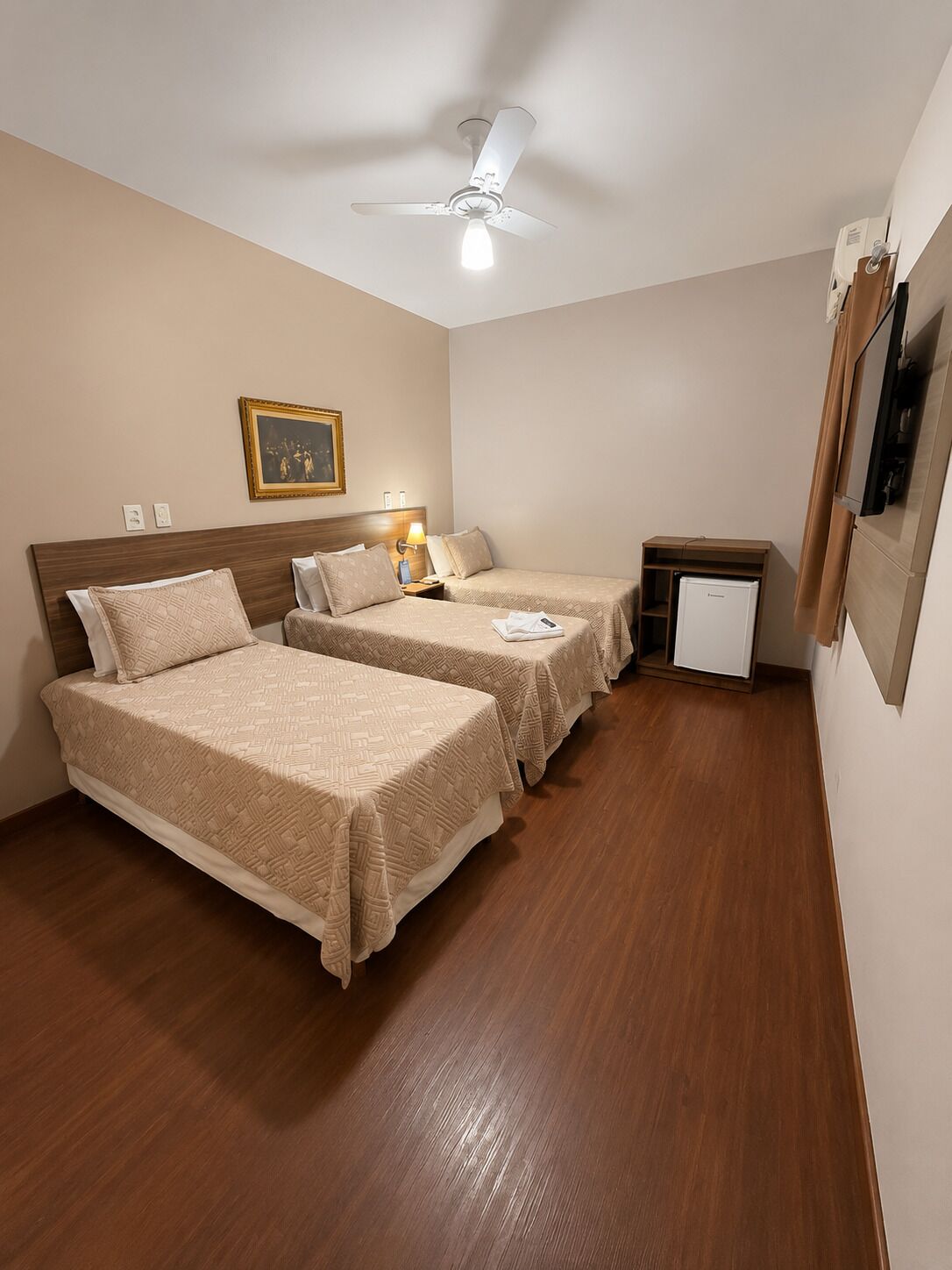 Habitación de hotel con dos camas, un ventilador de techo, un escritorio pequeño con refrigerador y una televisión montada en la pared.