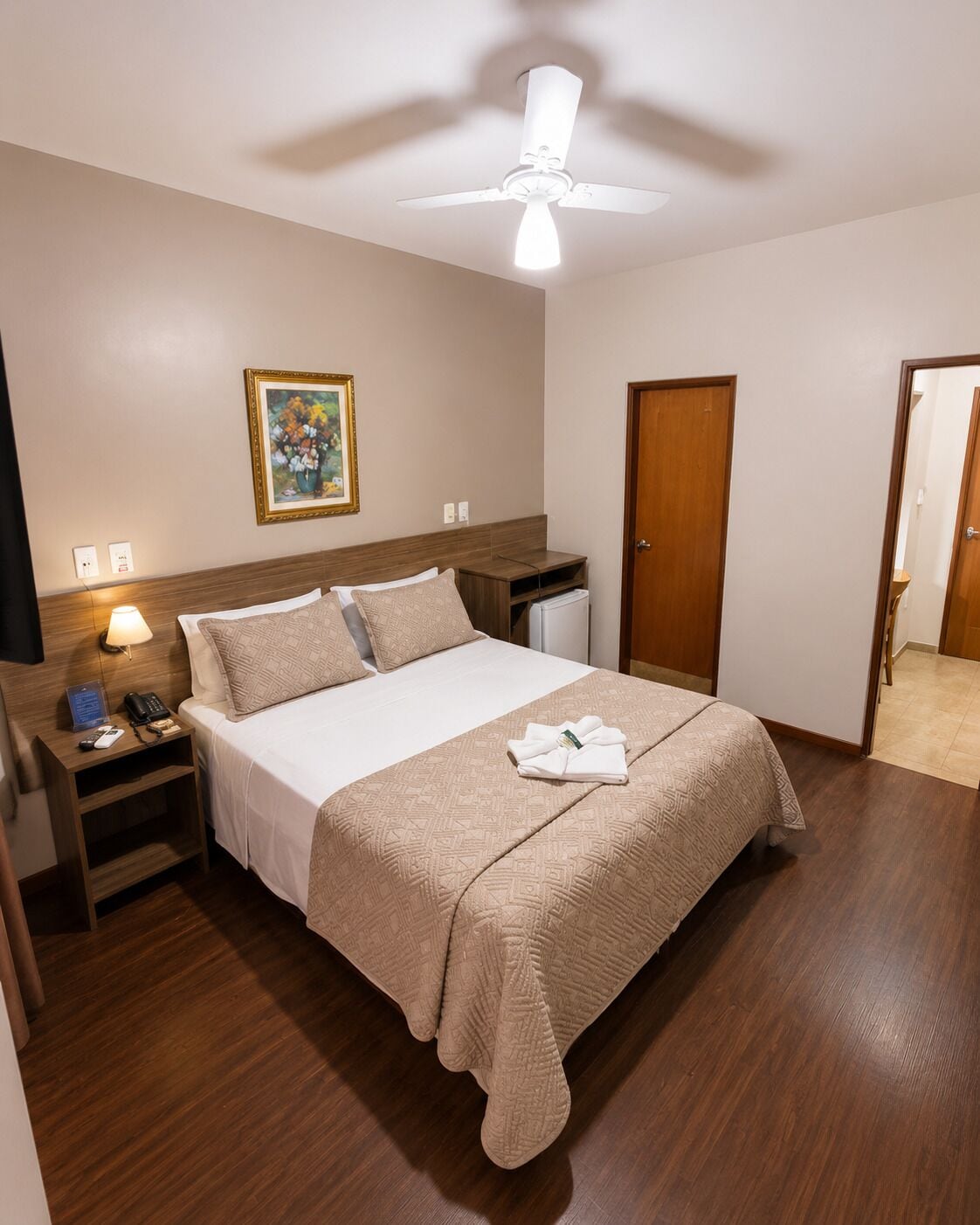 A hotel room with a bed, bedside table, lamp, ceiling fan, and a door.