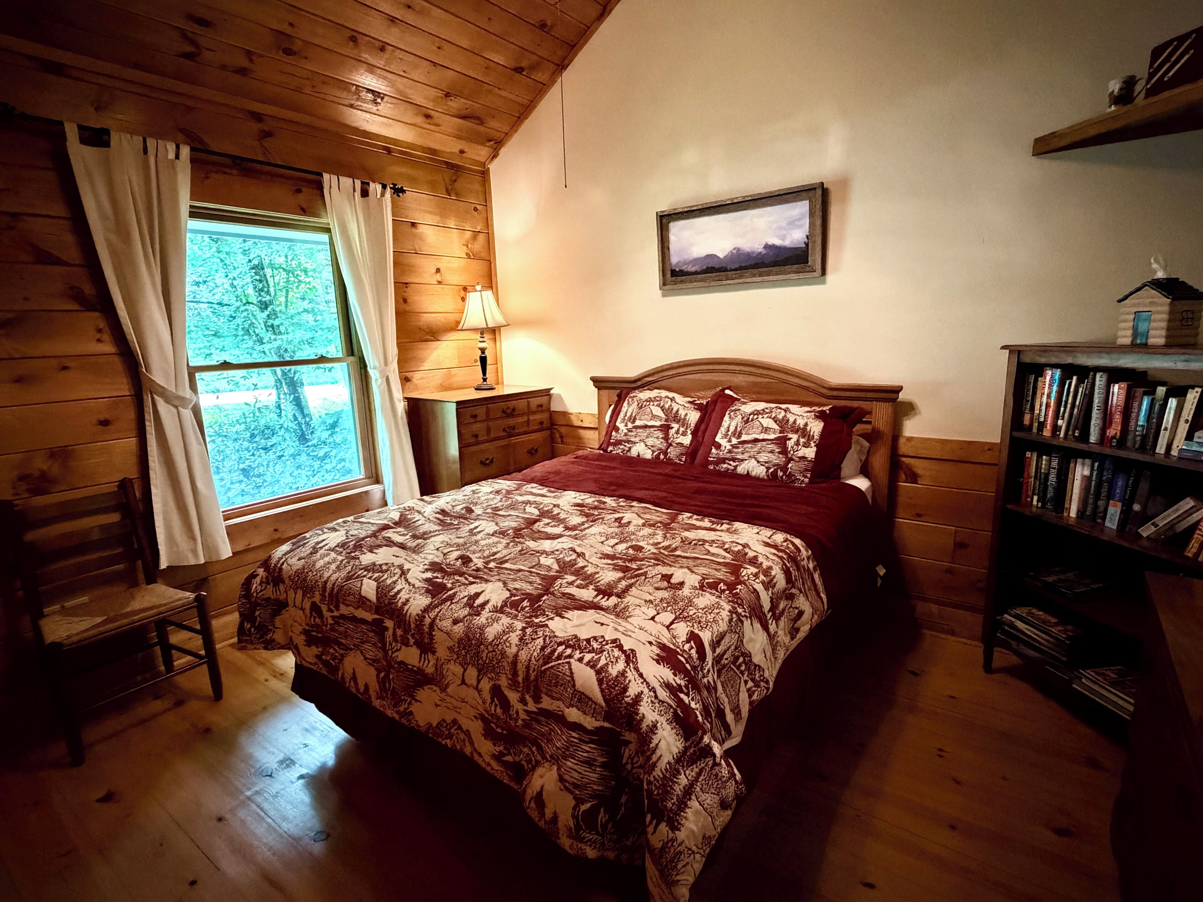 A wooden-paneled room with a bed, a nightstand, a lamp, a bookshelf, and a window with curtains.