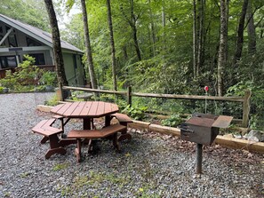A wooden picnic table with benches, a barbecue grill, and a fenced area with dense foliage. - Escape to the Cabin Between the Creeks a 2-bedroom house in Sugar Mountain, NC (Sugar Mountain)