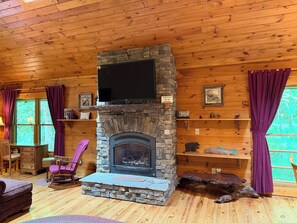 A cozy cabin interior with a stone fireplace, wooden walls, a television mounted above it, and purple curtains. - Escape to the Cabin Between the Creeks a 2-bedroom house in Sugar Mountain, NC (Sugar Mountain)