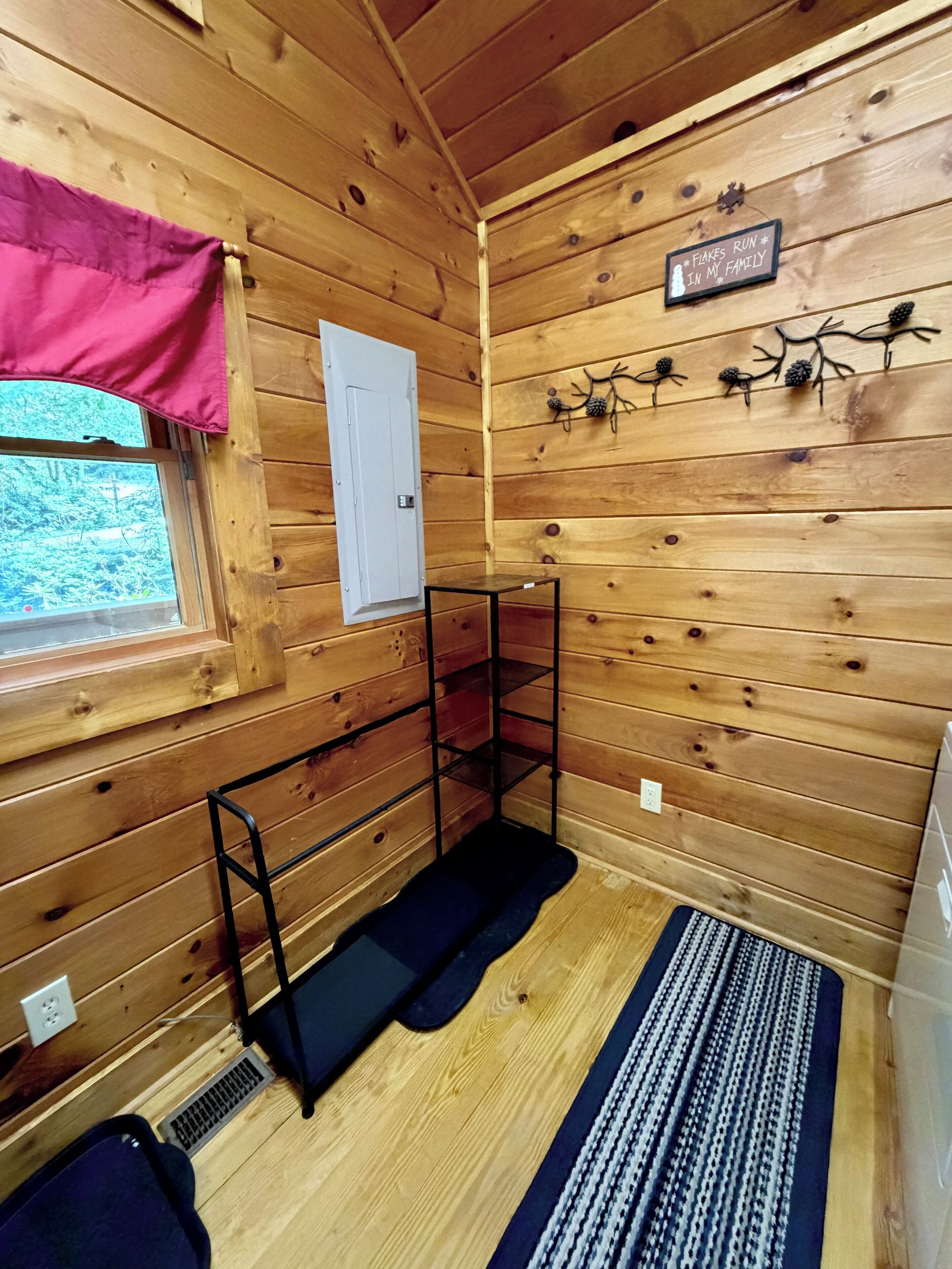 A wooden cabin interior with a window, a door, a metal shelf, a black mat, and a wall-mounted sign.