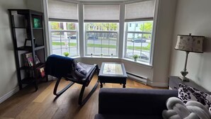 Una habitación con una mecedora, una mesita, una estantería y una ventana con vista a la calle.