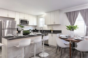 A modern kitchen with a central island, stainless steel appliances, white cabinetry, and a dining area with white chairs. - Chic City Apt Near Americana Glendale (Glendale)
