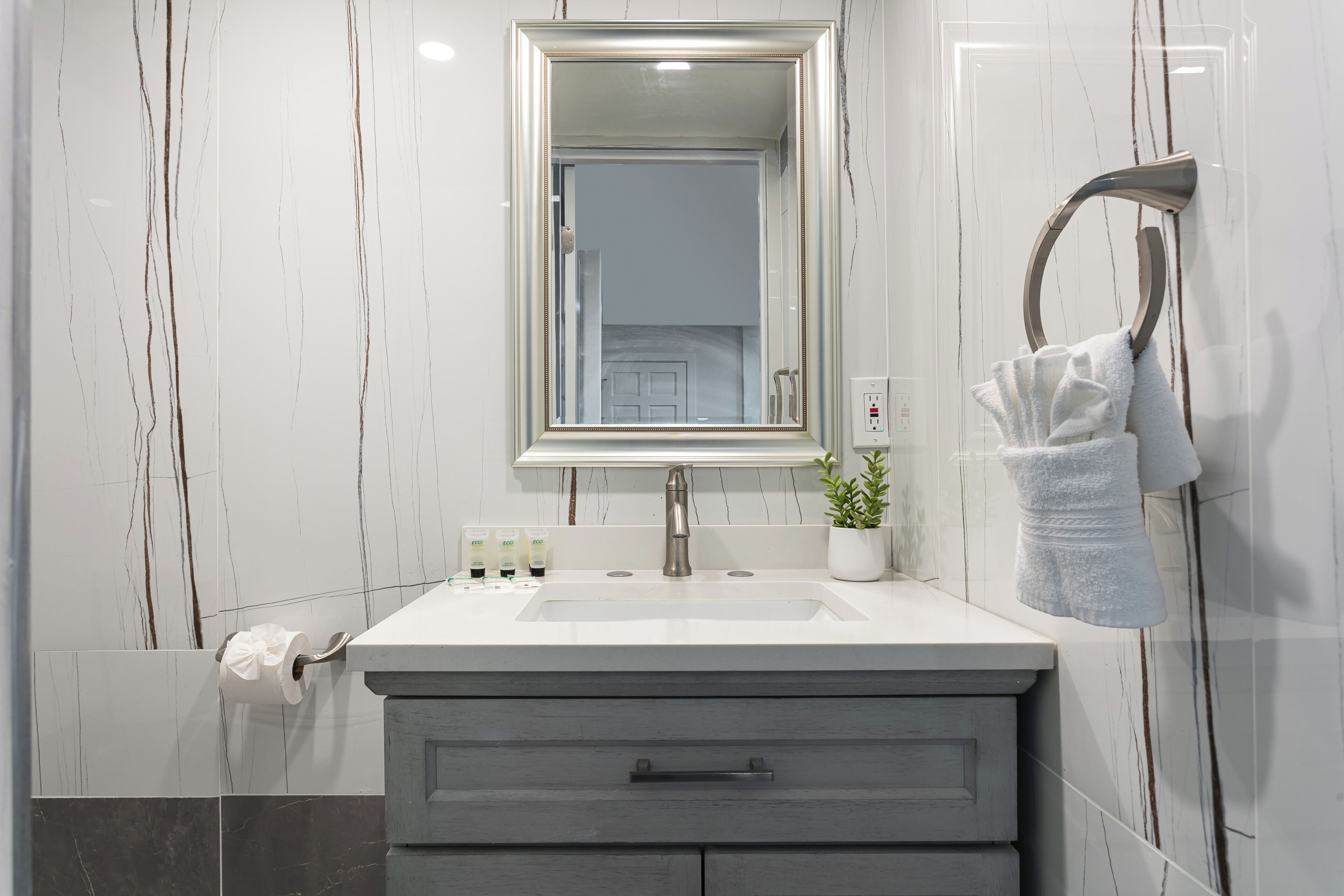 A bathroom with a marble wall, a sink with a mirror, a towel rack, and a plant.