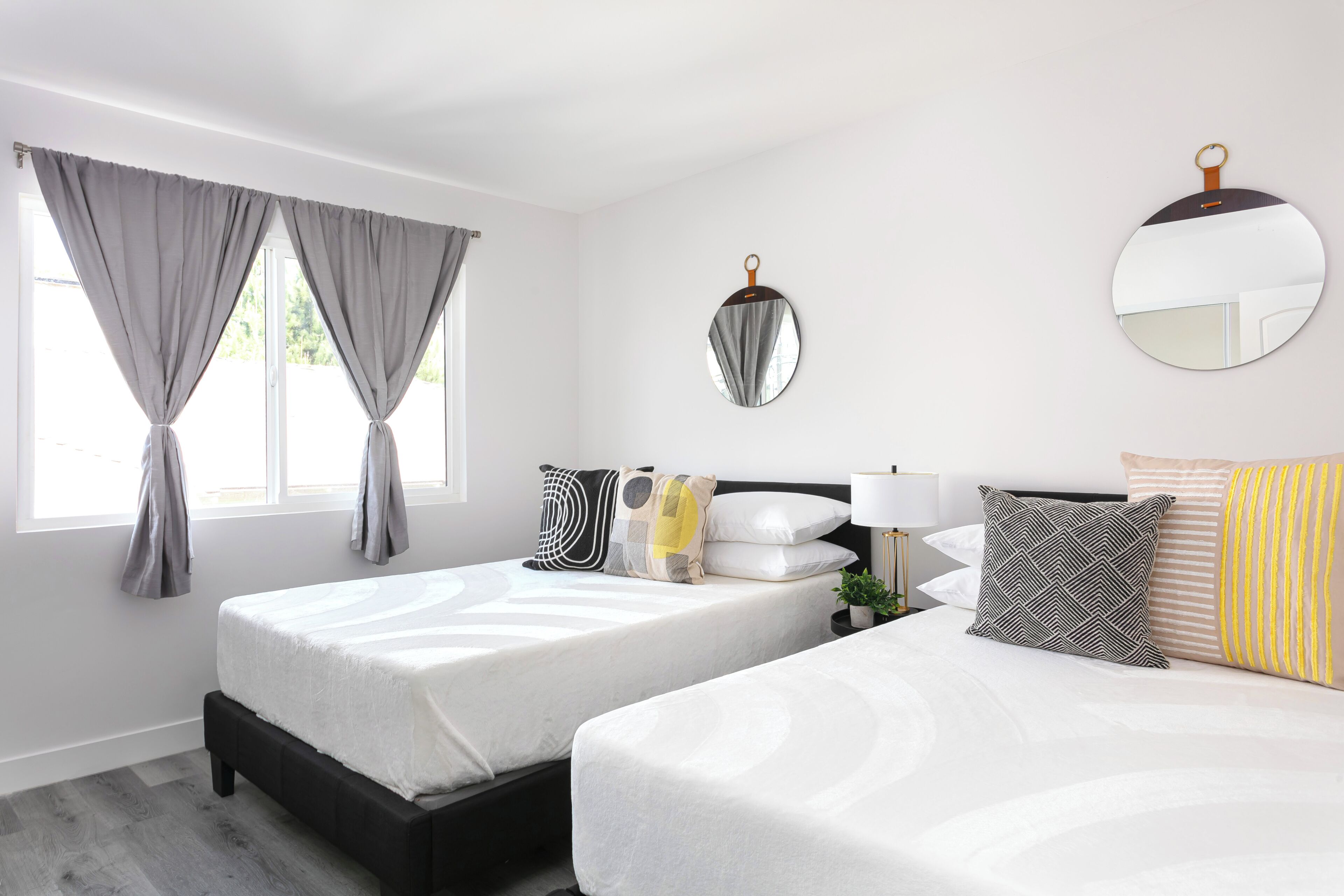 Un dormitorio con dos camas, un espejo redondo y un ventanal con cortinas.