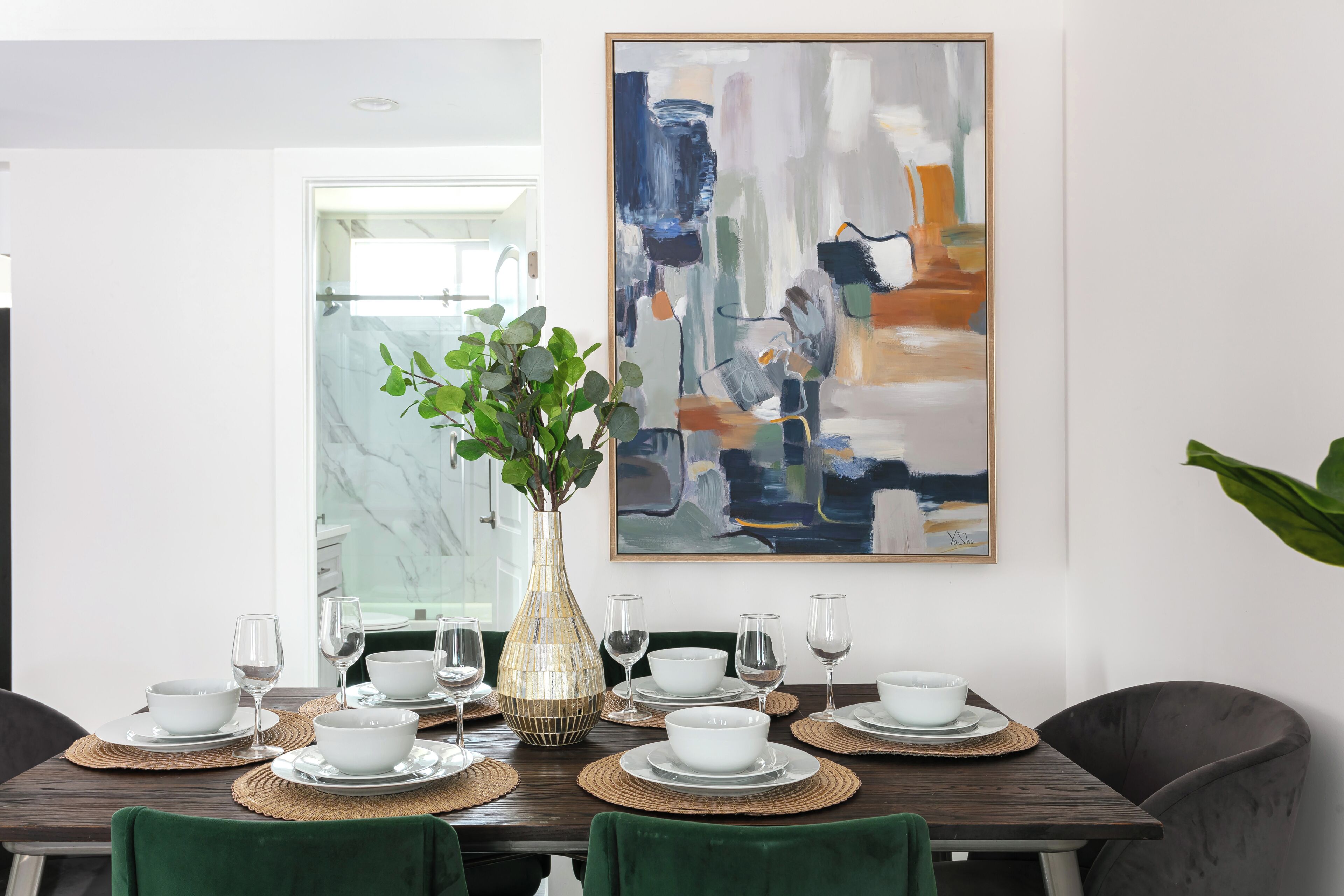 Una mesa de comedor dispuesta con cuencos y vasos blancos, un jarrón decorativo y un cuadro abstracto moderno en la pared.