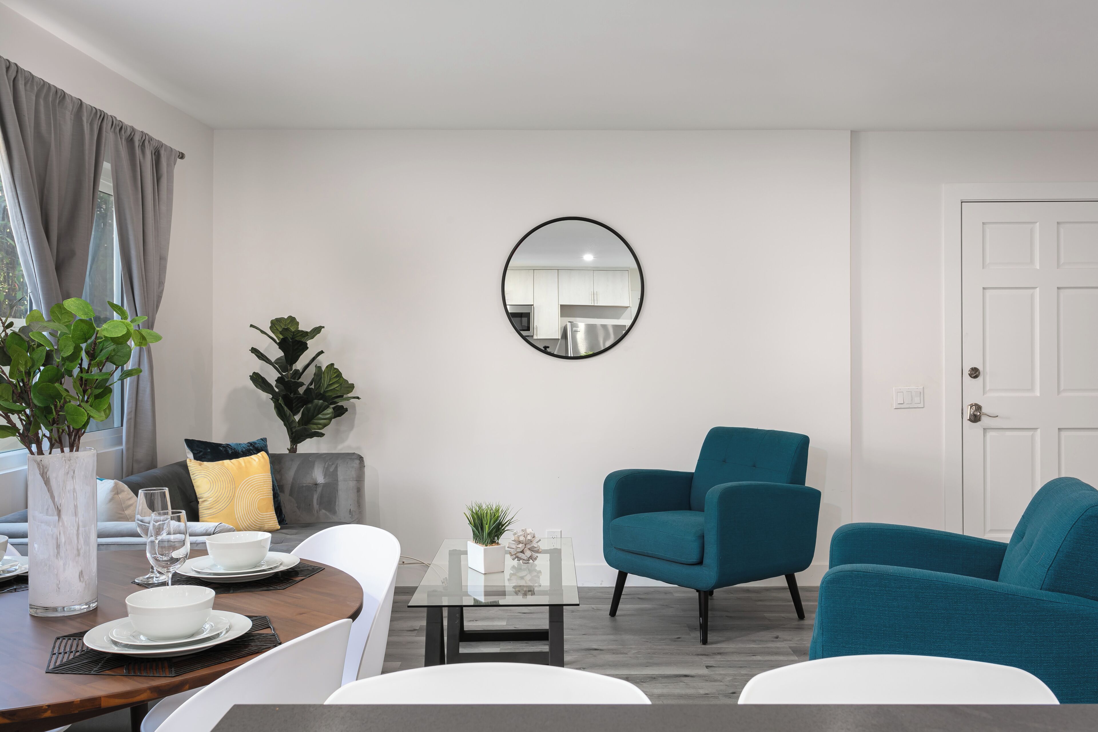 A modern living room with a dining area, a round mirror on the wall, and a view of the kitchen through the window.