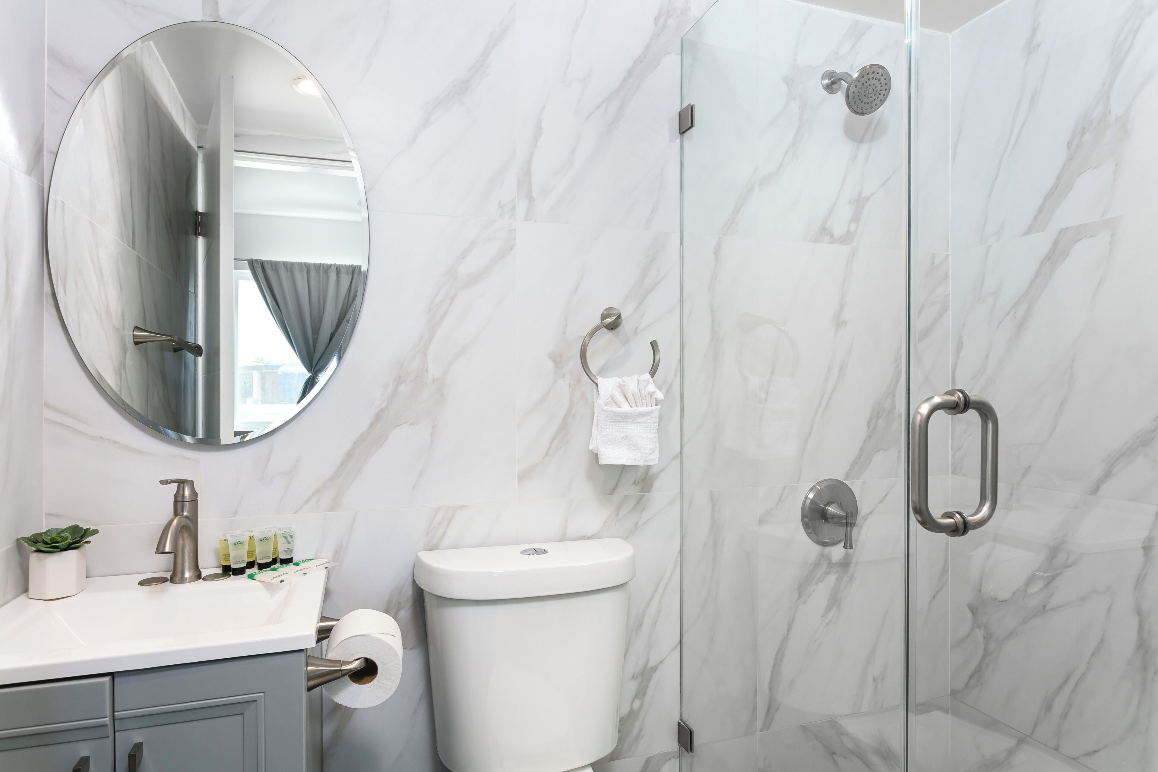 Un baño con pared de mármol, un espejo redondo, un inodoro, cabina de ducha de cristal y lavamanos con artículos de aseo.