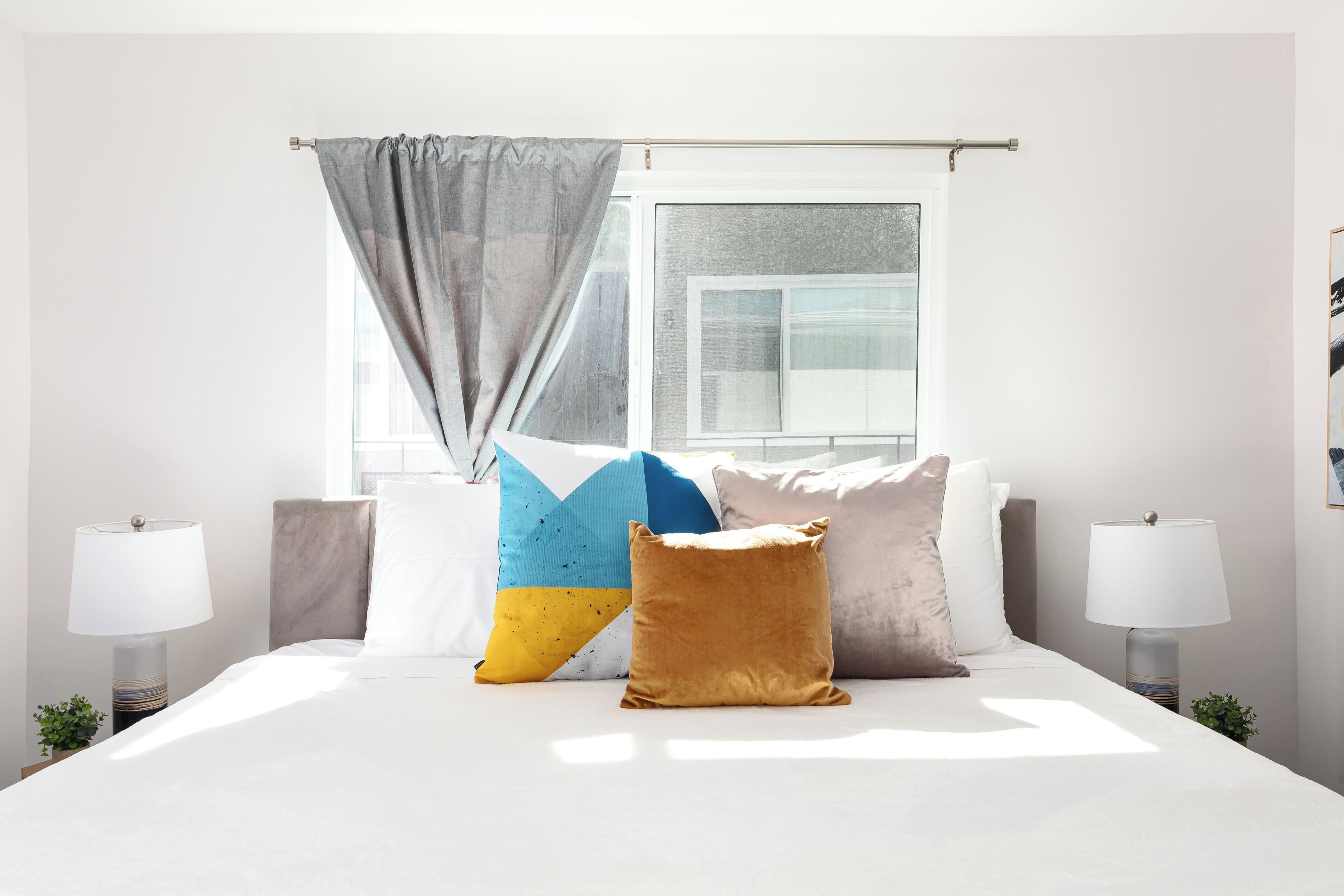 A hotel room with a bed, two bedside lamps, a window with curtains, and a view of buildings outside.
