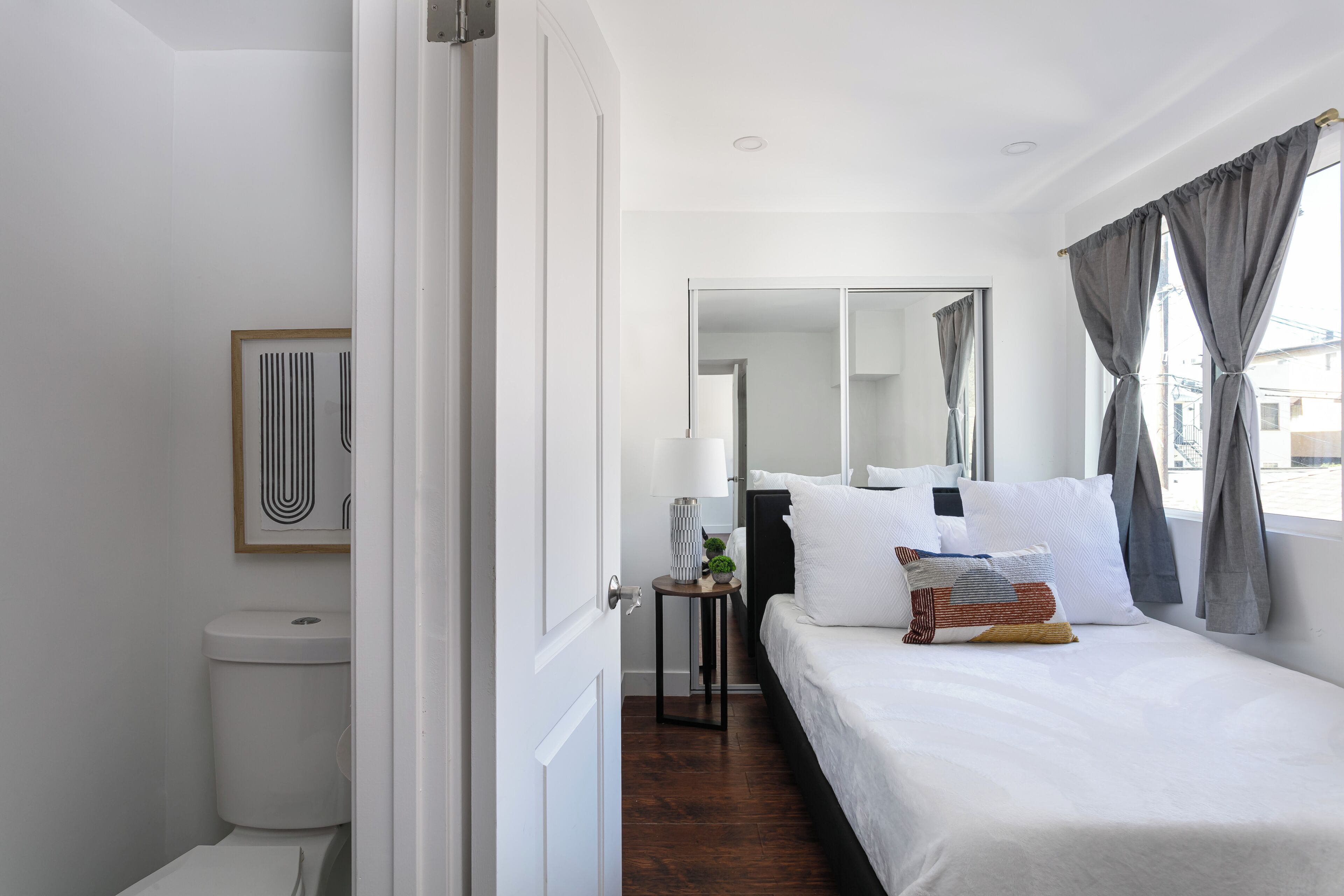 A hotel room with a bed, a bedside table, a mirror, a window with curtains, and a toilet.