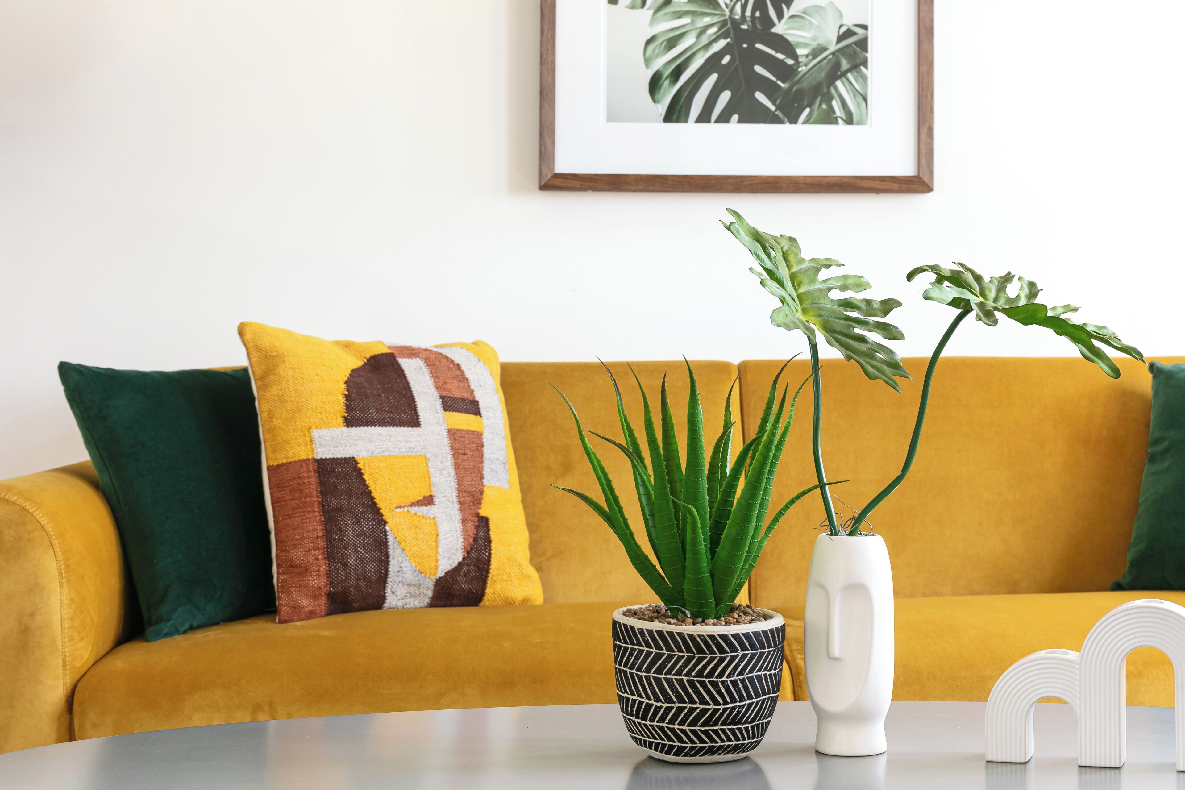 Un sofá amarillo con cojines estampados, una planta en maceta y un cuadro en la pared.