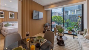 Une chambre d’hôtel moderne comprenant un lit, un coin repas avec une table et des chaises, un canapé, un téléviseur fixé au mur et de grandes fenêtres offrant une vue sur la ville.