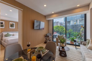 A modern hotel room with a bed, a dining area with a table and chairs, a sofa, a TV mounted on the wall, and large windows offering a city view.