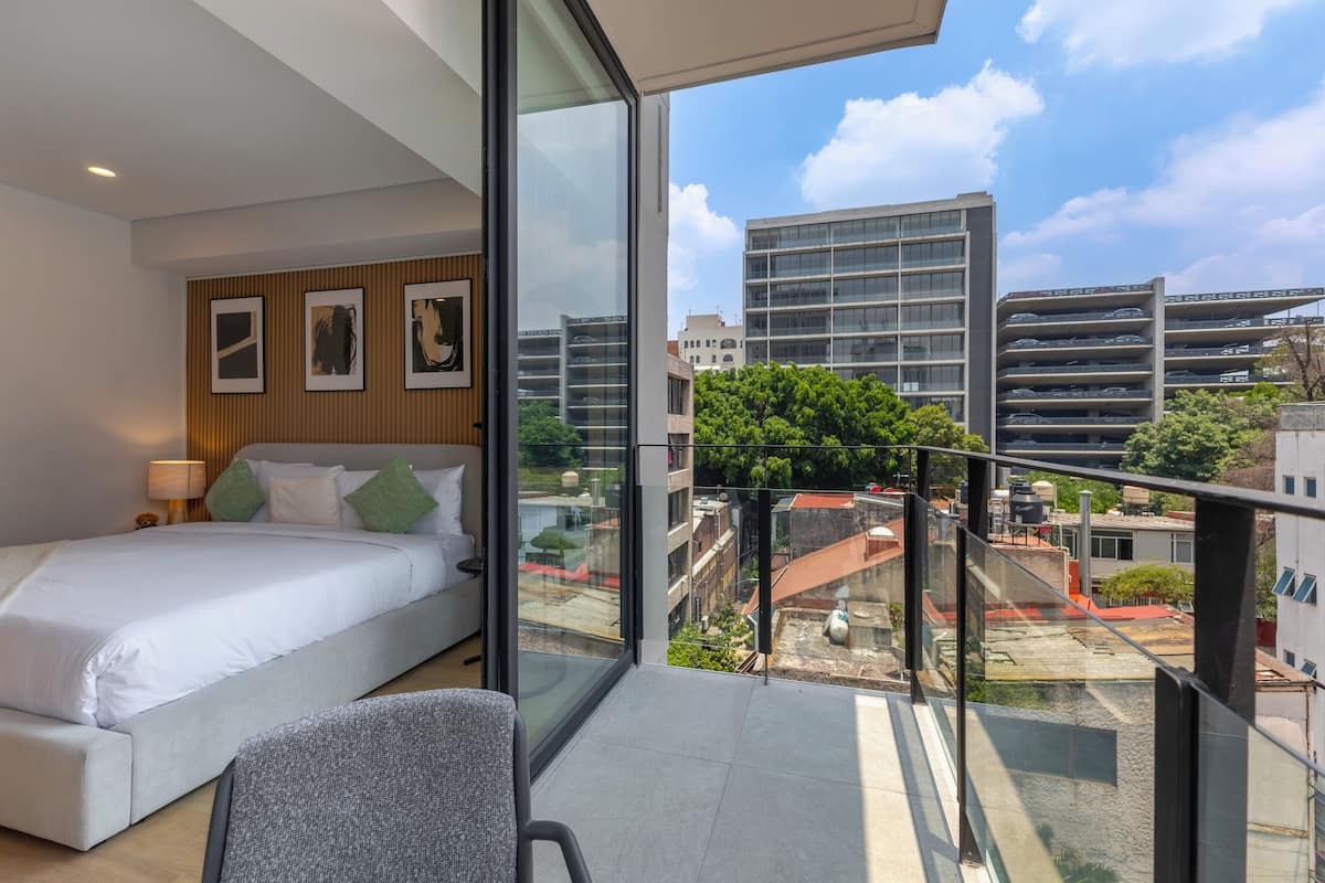 A modern hotel room with a large bed, a glass balcony, and a city view.