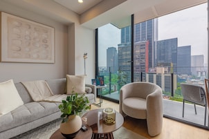 Uma sala de estar moderna com um sofá cinzento, uma mesa de centro redonda, uma poltrona cinzenta e uma janela grande com vista para a cidade.