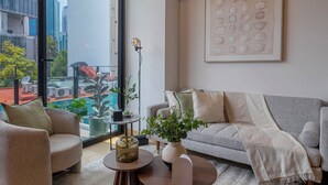 Un salon moderne avec un canapé, un fauteuil et une table basse, de grandes fenêtres offrant une vue sur la ville, et une œuvre d’art abstraite encadrée accrochée au mur.