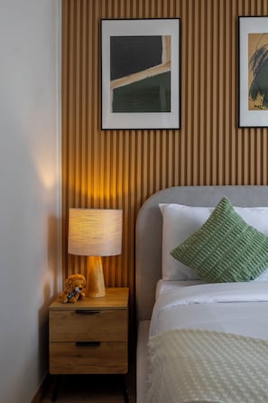 A neatly made bed with a green pillow, a bedside lamp, and a small teddy bear on a wooden nightstand.