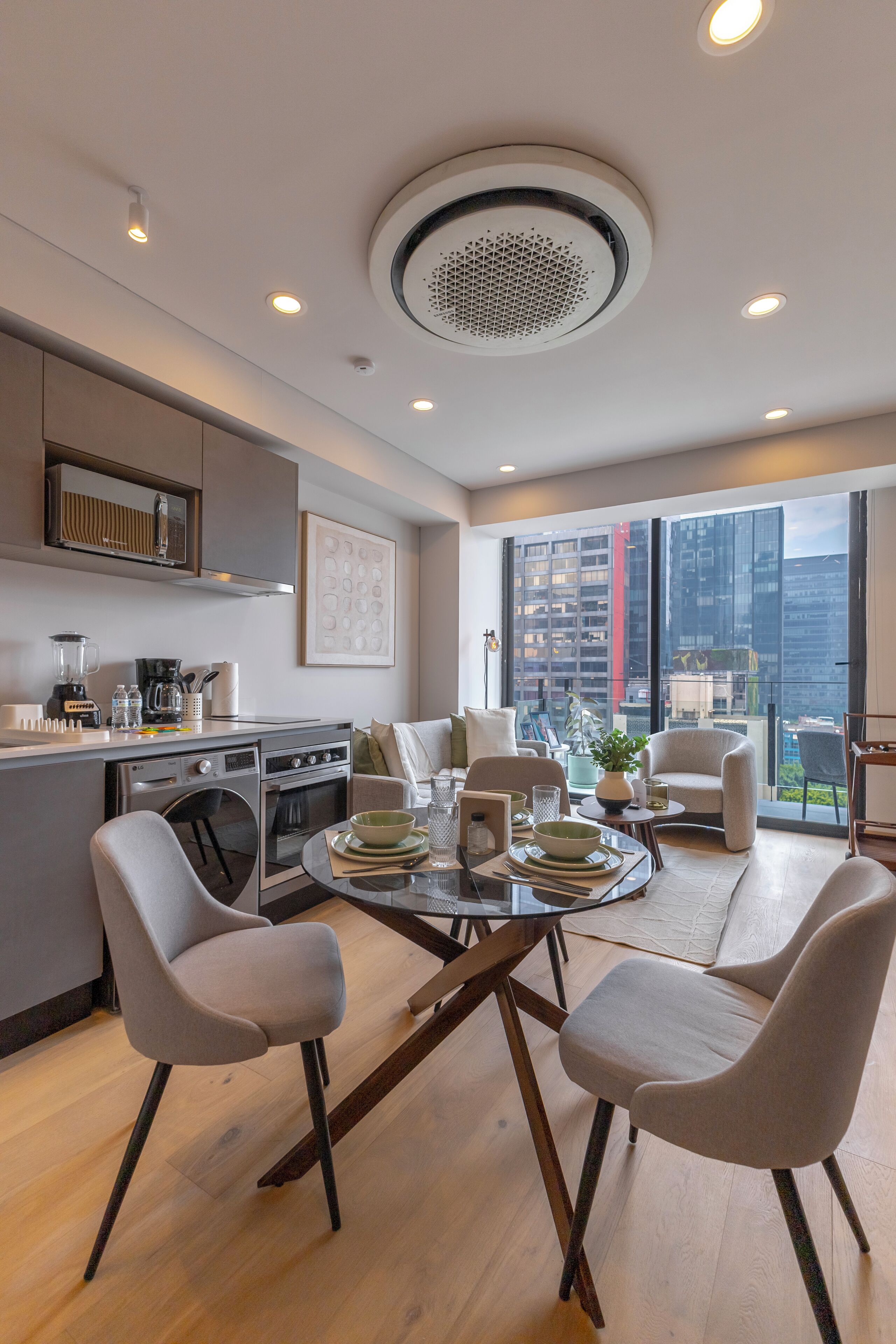Una cocina moderna con electrodomésticos integrados, un comedor con mesa de vidrio y vistas a un paisaje urbano a través de amplios ventanales.