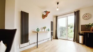 A modern living room with a wooden floor, a black chair, a radiator, a table with decorative items, a wall-mounted clock, and a large window with curtains. - super king size bed room with cozy and big bed for 3 in fmaily home (Hornchurch)