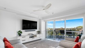 A modern living room with a flat-screen TV, a ceiling fan, a balcony with a table and chairs, and a city view. - The Surf Breeze Retreat (Coolangatta)