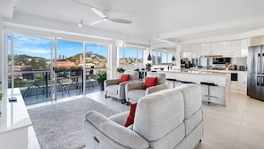 A modern living room with a large window, a ceiling fan, and a kitchen area with stainless steel appliances. - The Surf Breeze Retreat (Coolangatta)